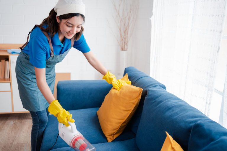 A woman wearing a blue shirt and yellow gloves is cleaning a blue couch with a cloth.